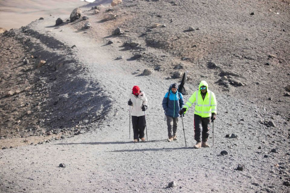 三名徒步者穿着分层装备走在火山岩小径上，地形与乞力马扎罗相似。