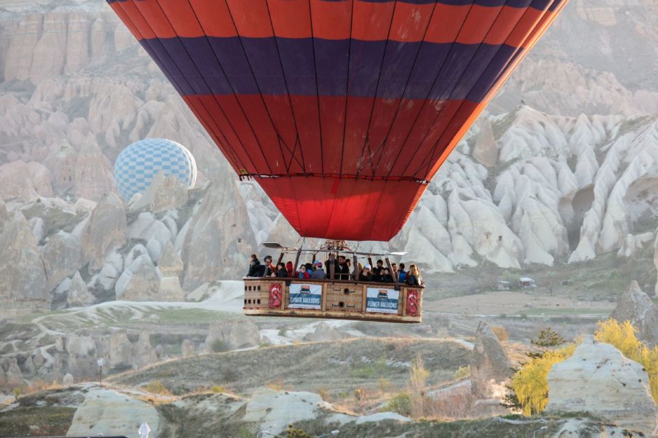 A hot-air balloon flies low over the rock formations of Cappadocia at sunrise, with another balloon behind.