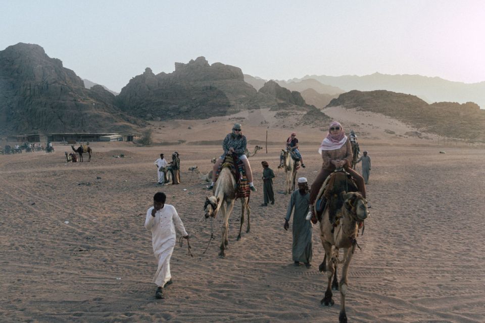 Visitors ride camels led by guides across the desert near Hurghada, with mountains behind.
