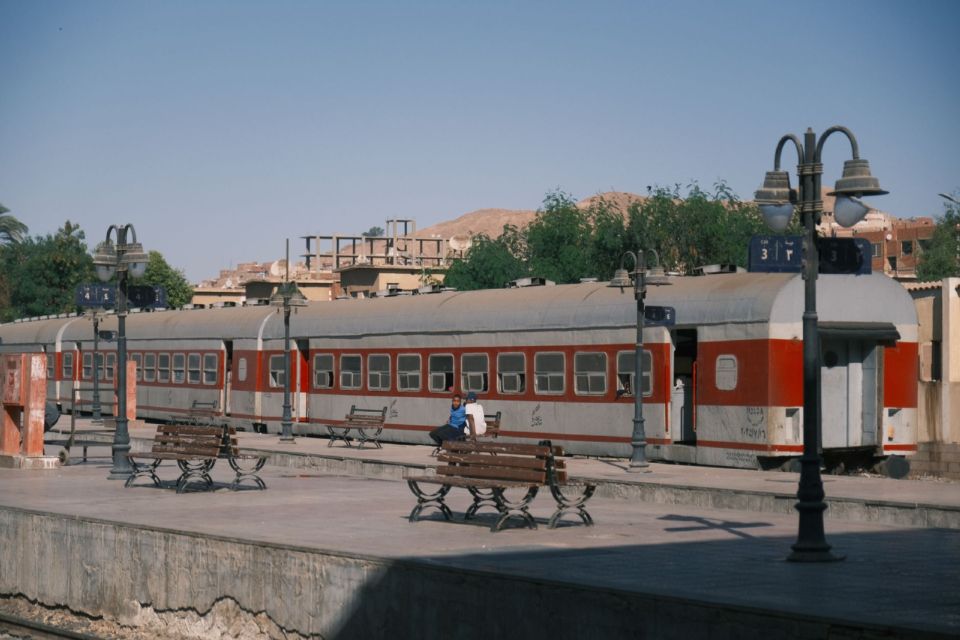 A train waits at a platform in Aswan, with hills and palm trees in the background.